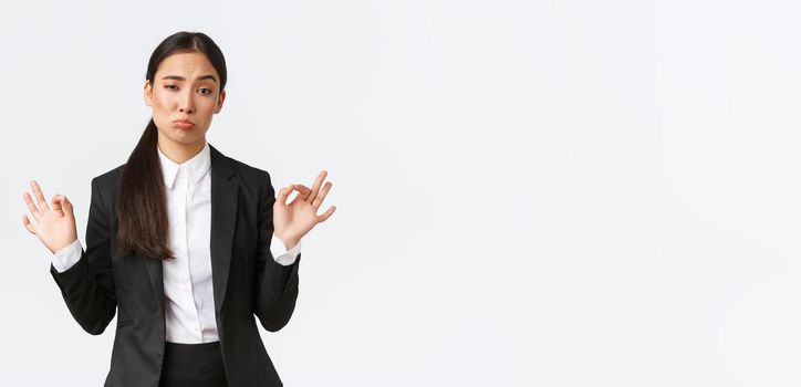 Pleased sassy asian businesswoman in black suit show not bad gesture, nod in approval and make okay gesture, very good work, praising coworker, standing satisfied over white background.