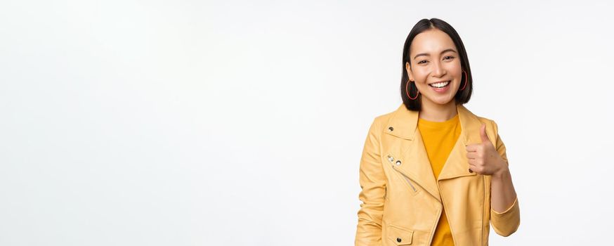 Beautiful korean girl smiling, showing thumbs up, like gesture, recommending store or company, standing satisfied against white background.