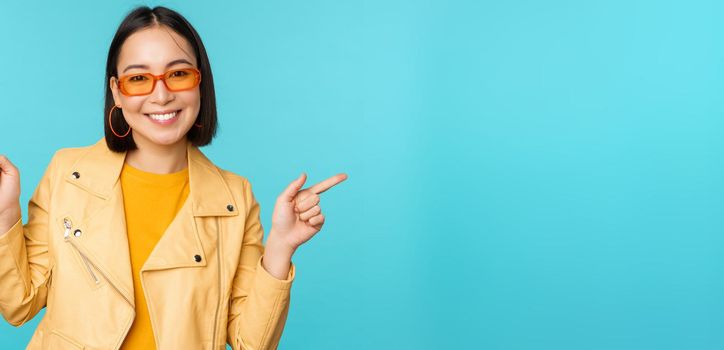 Happy stylish chinese girl in sunglasses, points fingers sideways, invites to check out, see store discount, shows left and right, stands over blue background.