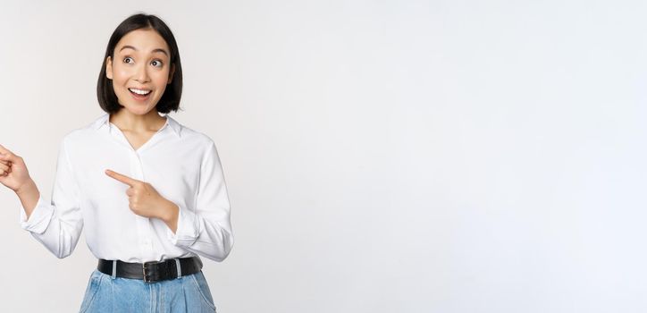 Enthusiastic korean girl pointing fingers left, female student pointing and looking left with happy smile, showing company logo or banner, white background.