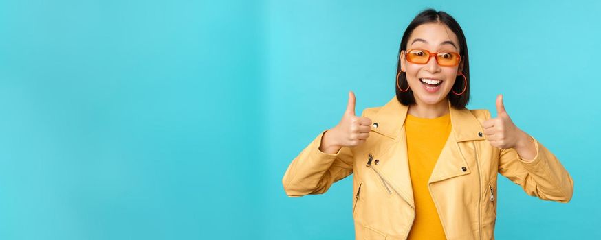 Enthusiastic korean girl looks excited and shows thumbs up, approves smth awesome, excellent choice and quality, stands over blue background.