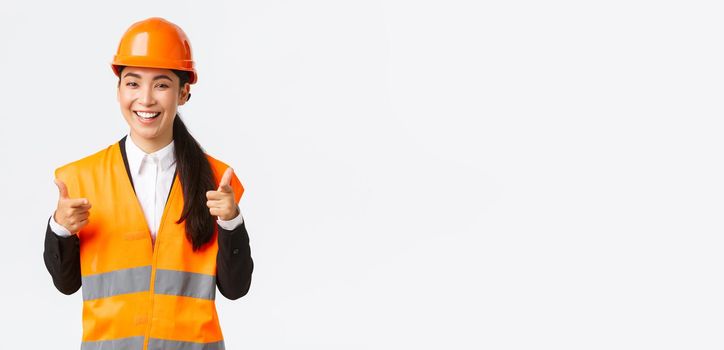 Confident pretty asian female construction manager in safety helmet and reflective jacket, pointing fingers at camera praising nice choice, good work or well done, standing white background.