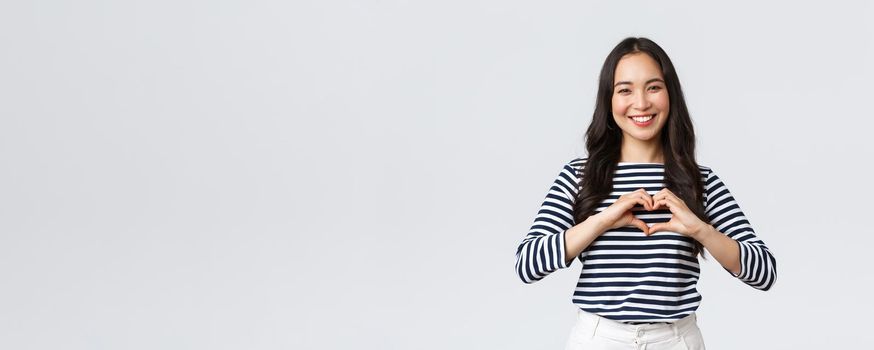 Lifestyle, people emotions and casual concept. Lovely smiling adorable asian woman showing heart sign and smiling, express sympathy or care, standing white background.