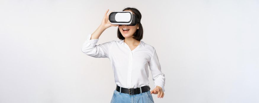 Excited young woman enter virtual reality in her glasses. Asian girl using vr headset, standing over white background.