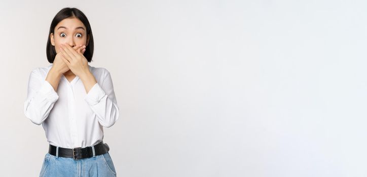 Shocked young asian girl close mouth, cover lips with hands and looking amazed, standing over white background. Taboo gesture