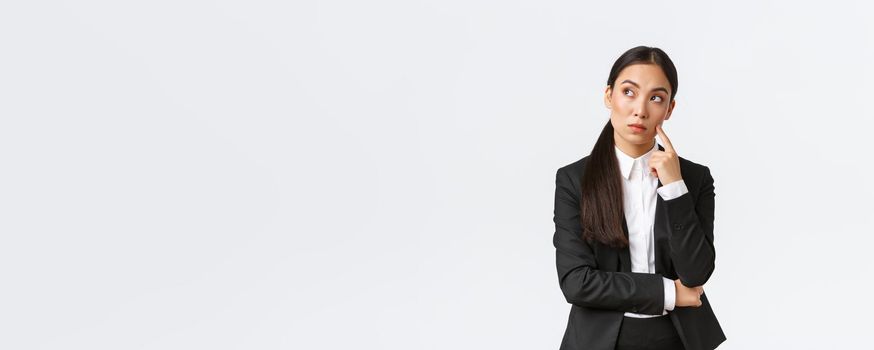 Thoughtful serious-looking asian female manager, office worker in suit thinking about solution, making hard choice, looking away while pondering, standing white background.