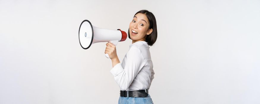 Beautiful young asian woman talking in megaphone, screams in speakerphone and smiling, making announcement, shout out information, standing over white background.