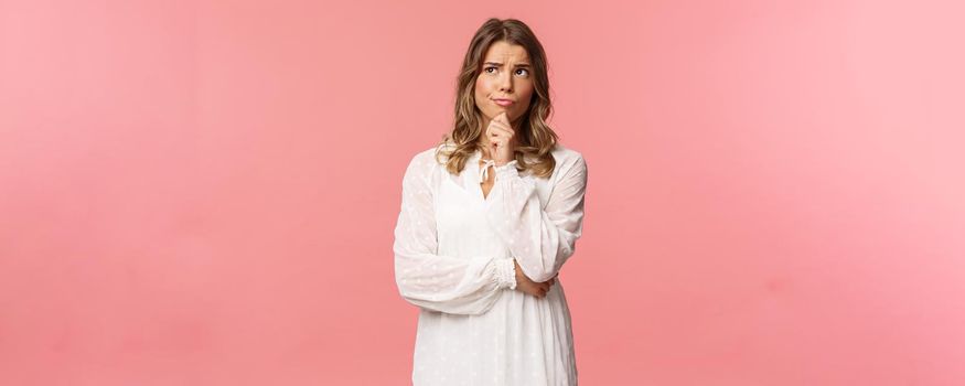 Beauty, fashion and women concept. Portrait of uncertain, serious-looking feminine young blond woman in white dress, touch chin, smirk and frowning thoughtful, look away thinking.