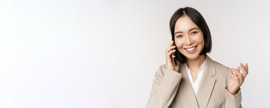 Smiling corporate woman in suit, talking on mobile phone, having a business call on smartphone, standing over white background.