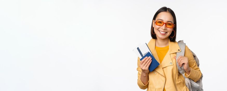 Happy asian girl going on vacation, holding passport and flight tickets, backpack on shoulder. Young woman tourist travelling abroad, standing over white background.