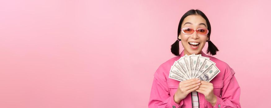 Microcredit and fast loans concept. Excited stylish korean girl, showing money, cash dollars and looking happy, standing in sunglasses over pink background.