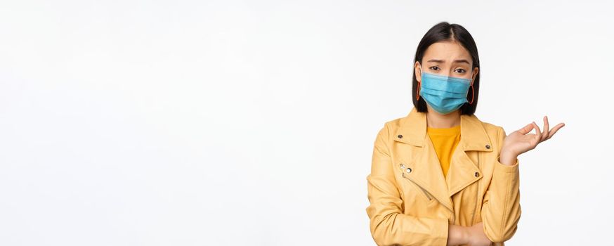 Confused asian woman in medical face mask, looking clueless, puzzled, wearing medical face mask, standing over white studio background.