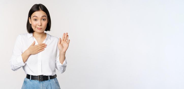 Image of cute young female office worker, asian girl student raising hand up and put palm on chest, name herself, introduce, making promise, standing over white background.