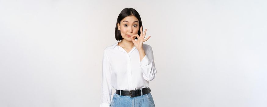 Mouth shut. Young asian woman seal lips, showing taboo dont speak, keep quiet sign, hiding secret, standing over white background.