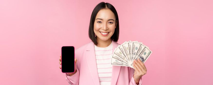 Image of korean successful corporate woman showing money, dollars and smartphone app screen, interface of mobile phone application, concept of investment and finance, pink background.