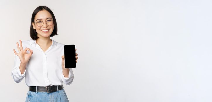 Image of asian businesswoman, showing smartphone screen, app interface and ok sign, recommending application on mobile phone, standing over white background.