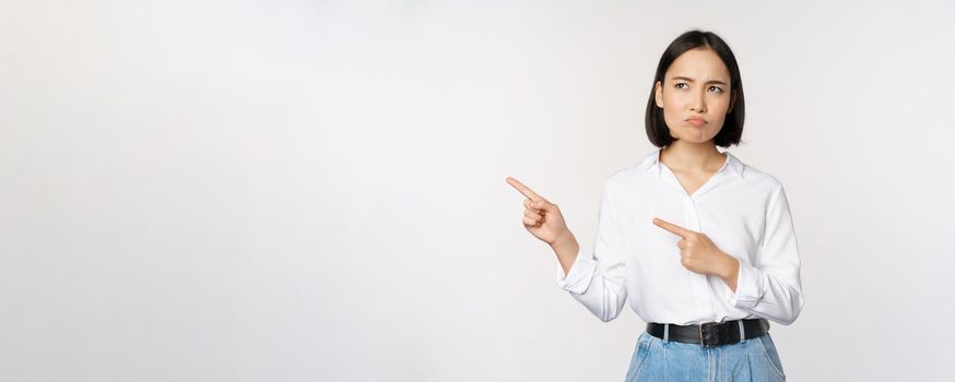 Disappointed, sad young asian woman pointing and looking left with upset, sulking face expression, standing over white background.