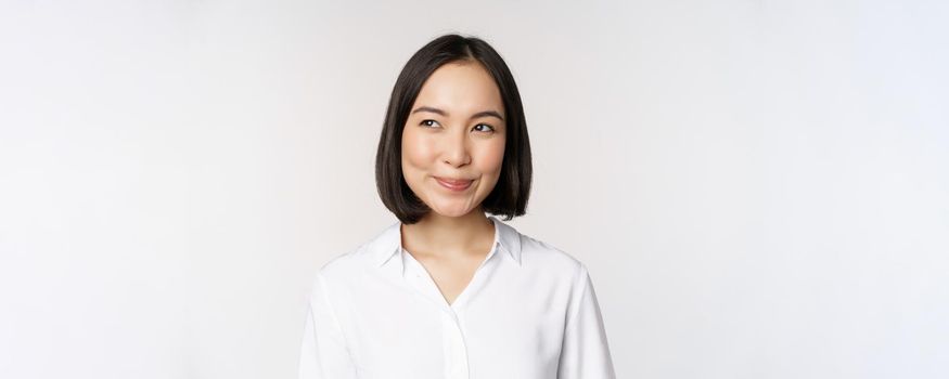 Image of smiling asian woman planning, thinking of smth, daydreaming, standing over white background with smug face. Copy space