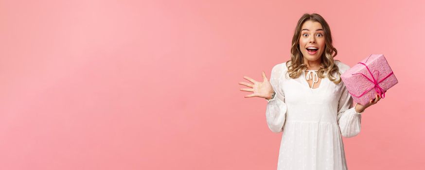 Holidays, celebration and women concept. Portrait of surprised upbeat charismatic blond girl in white dress, spread hands sideways and gasping amazed, receive surprise gift, hold pink box.