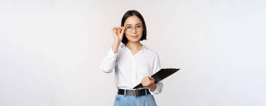 Young woman, office worker manager in glasses, holding clipboard and looking like professional, standing against white background.