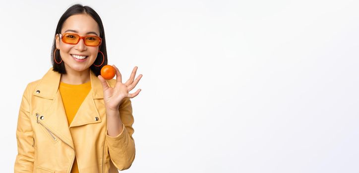 Beautiful asian girl in sunglasses showing tangerine and smiling, looking happy, posing in yellow against studio background.