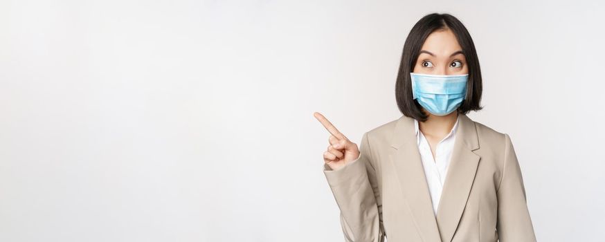 Enthusiastic businesswoman pointing fingers left, wearing medical face mask from covid-19 pandemic, standing over white background.