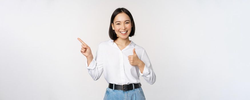 Image of young asian business woman, smiling while pointing finger left and showing thumbs up, recommending product, praise, standing over white background.