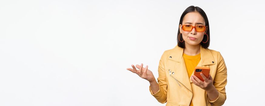 Portrait of confused asian girl in sunglasses, holding smartphone, using mobile phone and looking puzzled, standing clueless over white background.