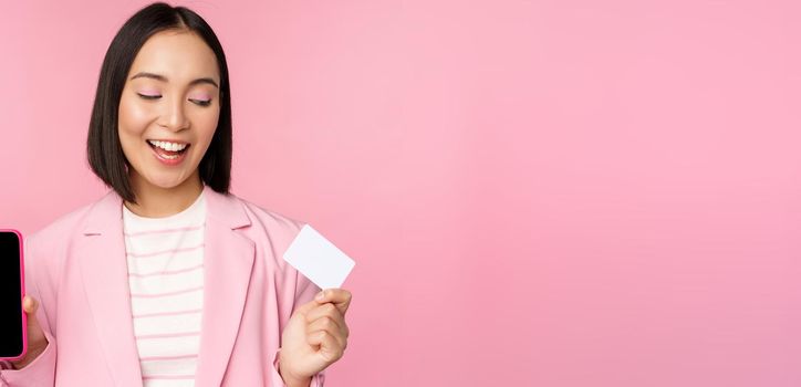 Smiling korean businesswoman in suit, showing mobile phone screen, credit card, showing online banking application interface, pink background.