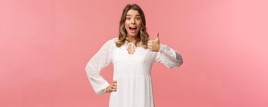 Great idea. Upbeat, enthusiastic blond caucasian girl in white spring dress, hear good plan, show thumbs-up and nod in approval, agree and recommend, satisfied with choice, stand pink background.
