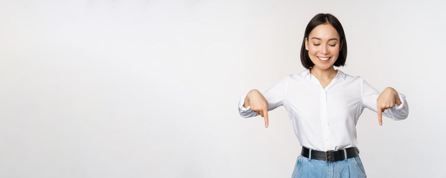 Portrait of happy asian woman pointing fingers down and looking below at advertisement, showing info banner, advertising, standing over white background.