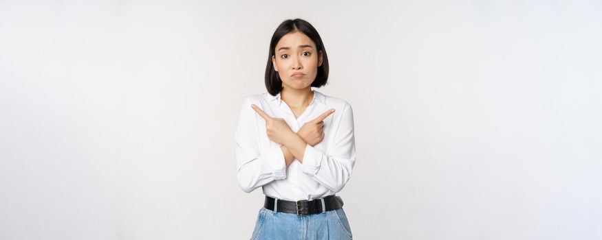 Confused sad asian girl cant decide, pointing fingers left and right, sulking and shrugging indecisive, cannot choose, standing over white background.