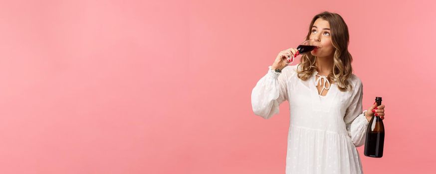 Holidays, spring and party concept. Portrait of young elegant happy blond woman, wear white trendy dress, drinking wine from glass looking up pleased, tasting good drink, hold bottle.