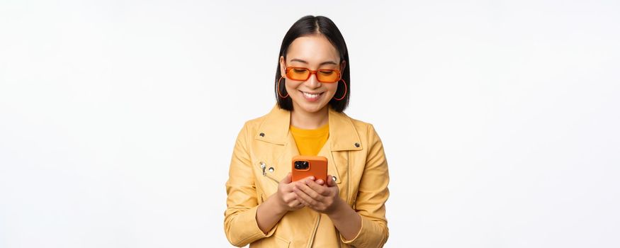 Modern asian girl in sunglasses using her mobile phone, smiling and looking happy, posing against white background. Copy space