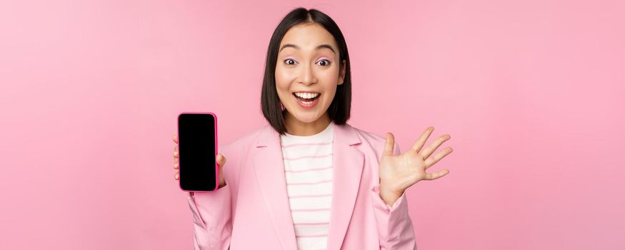 Surprised, enthusiastic asian businesswoman showing mobile phone screen, smartphone app interface, standing against pink background.