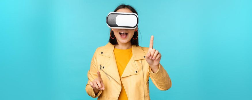 Young asian woman using virtual reality glasses, using VR headset, standing amused against blue background. Copy space