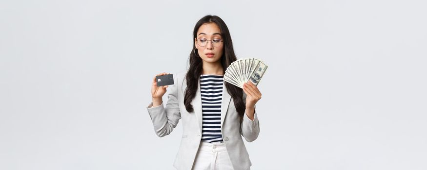 Business, finance and employment, entrepreneur and money concept. Indecisive cute asian office lady thinking about putting cash on deposit, looking curious at credit card, white background.