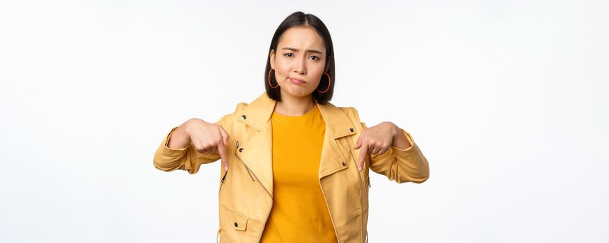 Portrait of disappointed asian woman pointing fingers down, grimacing and showing smth bad, standing over white background. Copy space