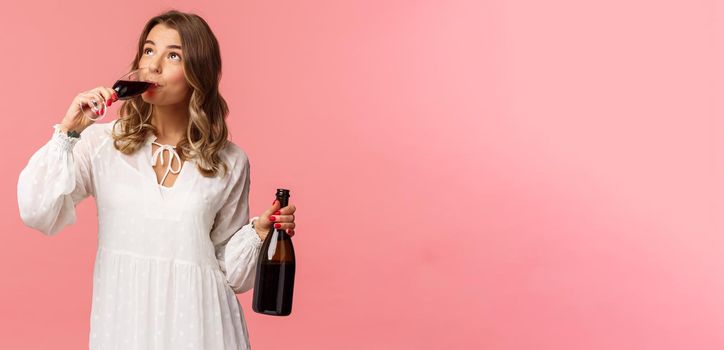 Holidays, spring and party concept. Portrait of young elegant happy blond woman, wear white trendy dress, drinking wine from glass looking up pleased, tasting good drink, hold bottle.
