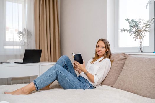 White cozy bed and a beautiful girl in a white shirt and jeans reading a book, the concept of home and comfort. Against the background of the living room, computer and window