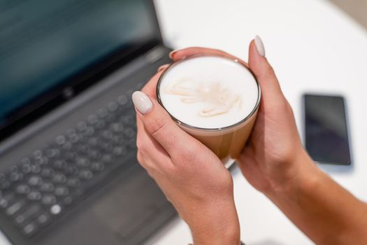 Close-up of beautiful female hands holding a large white cup of cappuccino. A business woman sits in a cafe working at a computer