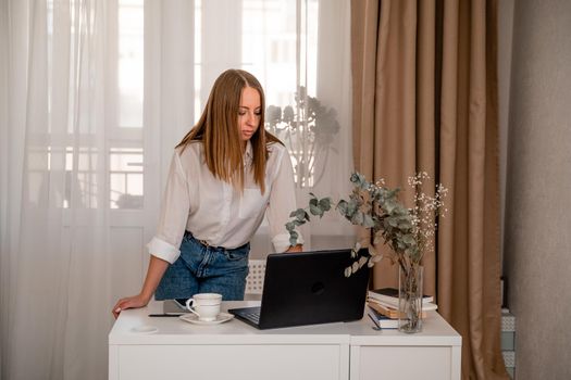 European professional woman is sitting with a laptop at a table in a home office, a positive woman is studying while working on a PC. She is wearing a beige jacket and jeans and is on the phone