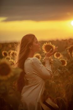 Beautiful middle aged woman looks good in a hat enjoying nature in a field of sunflowers at sunset. Summer. Attractive brunette with long healthy hair