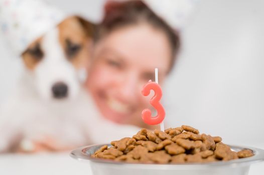 Caucasian woman and jack russell terrier in holiday caps look at a bowl of dry food with a candle. The dog and the owner are celebrating third birthday.