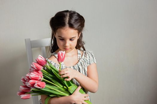 a beautiful romantic little girl with a bouquet of pink tulips, sits on a chair, against a gray wall. copy space