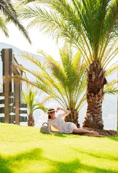 a bright beautiful girl in a light dress and hat lies on the grass under a palm tree in Monaco in sunny weather in summer, streets of old town of Monaco. High quality photo