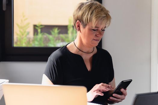 Smiling mature professional beautician with blonde hair, dressed in black work uniform, working in home office, looking at her mobile phone. Work from home, freelance, business, creative occupation, lifestyle concept. Relaxed atmosphere and soft lighting from window, natural light, work table, desk, computer. Horizontal.