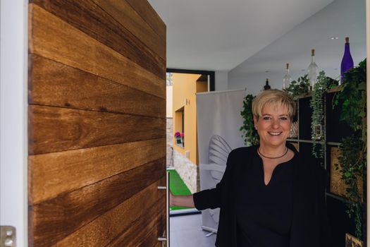 Mature, smiling professional beautician with blonde hair, dressed in black work uniform, greeting client in her small spa business. Business owner opening door to greet client. Work from home, freelance, business, creative occupation, lifestyle concept. Relaxed atmosphere and soft lighting from interior windows, natural light from outside, entrance door. Horizontal.