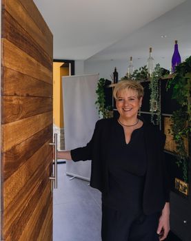 Mature, smiling professional beautician with blonde hair, dressed in black work uniform, greeting client in her small spa business. Business owner opening door to greet client. Work from home, freelance, business, creative occupation, lifestyle concept. Relaxed atmosphere and soft lighting from interior windows, natural light from outside, entrance door. Vertical