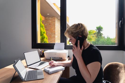 Mature and smiling professional beautician woman with blonde hair, dressed in black work uniform, working in her home office. Owner of a beauty salon receiving phone consultations. Work from home, freelance, business, creative occupation, lifestyle concept. Relaxed atmosphere and soft lighting from window, natural light, work table, desk, computer. Horizontal.